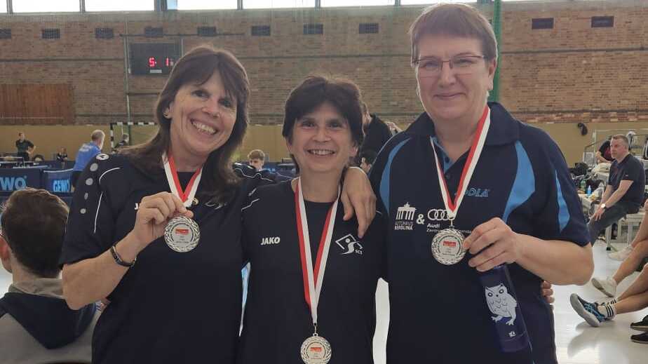 Drei Frauen in Sportkleidung halten Medaillen in einer Sporthalle, lächeln und posieren nebeneinander.