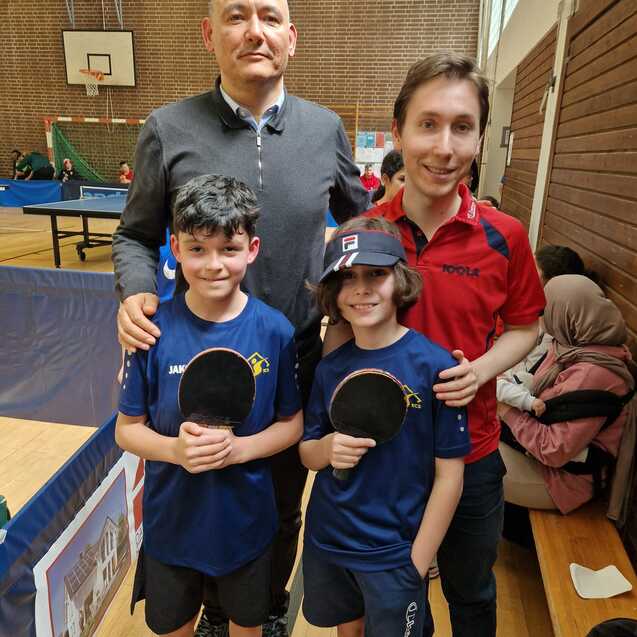 Vier Personen stehen in einer Sporthalle zusammen, zwei Kinder mit Tischtennisschlägern und zwei Erwachsene im Hintergrund.