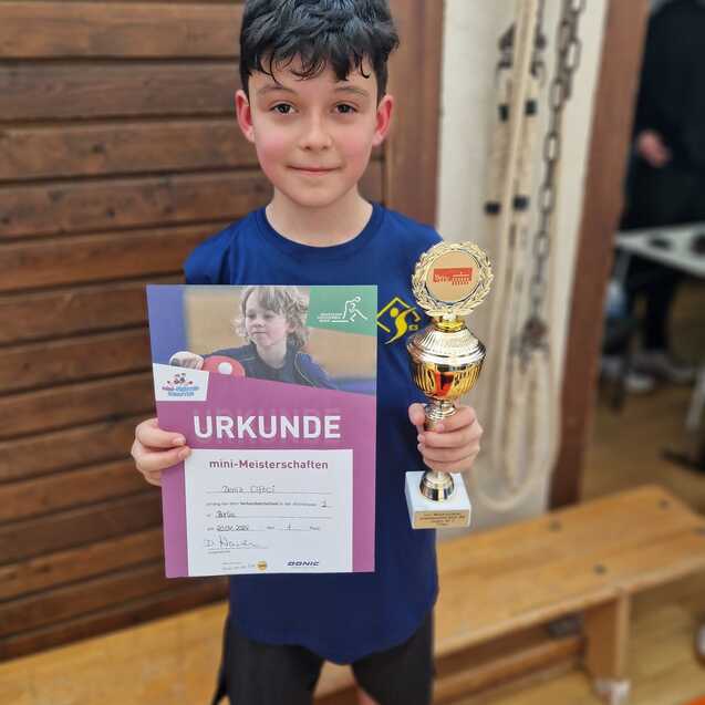Ein Junge hält eine Urkunde und einen Pokal nach den mini-Meisterschaften in einer Innenraumumgebung.