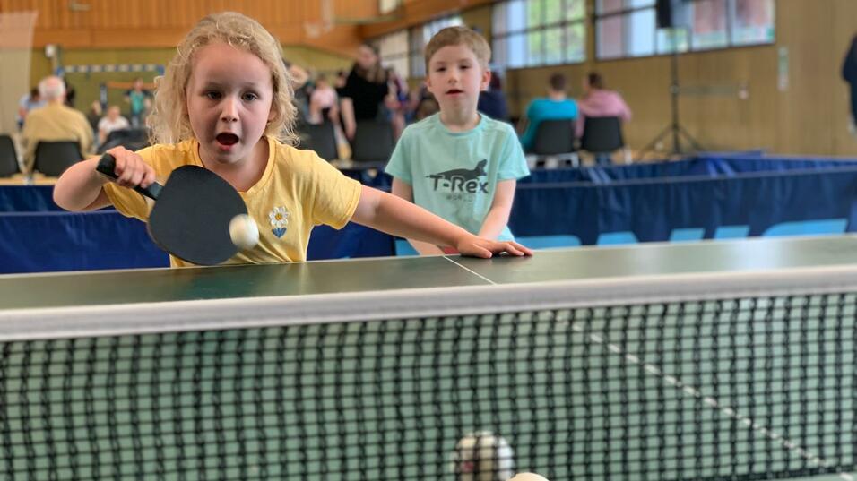 Ein Mädchen mit blonden Locken spielt Tischtennis, während ein Junge im Hintergrund zuschaut.