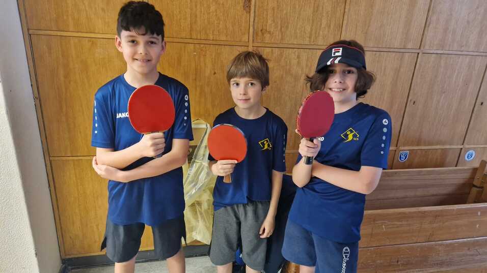 Drei Kinder in blauen Sportshirts halten Tischtennisschläger vor einer Holzwand und einer Bank.