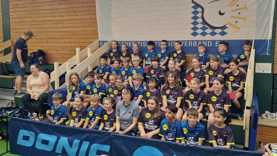 Gruppenfoto von Kindern in Sportkleidung mit Tischtennis-T-Shirts, versammelt vor einem Banner im Turnierumfeld.