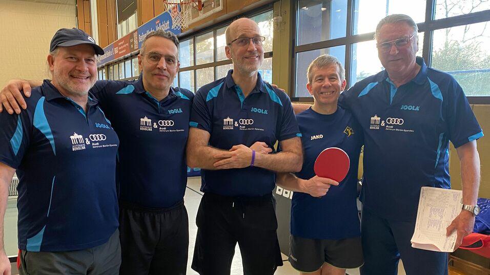 Fünf Tischtennisspieler in Uniformen stehen lächelnd in einer Sporthalle mit Tischtennisplatte im Hintergrund.