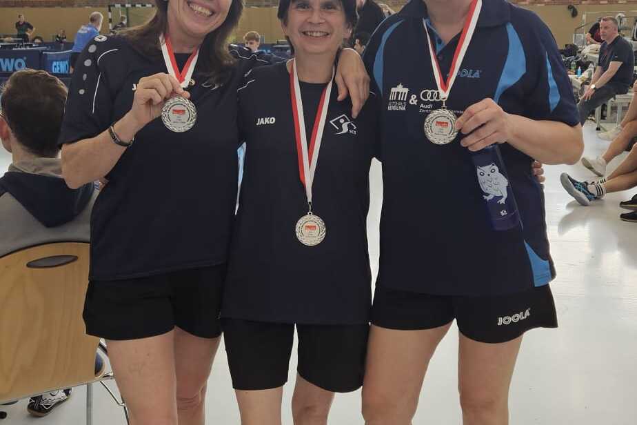 Drei Frauen in Sportkleidung halten Medaillen in einer Sporthalle, l&auml;cheln und posieren nebeneinander.