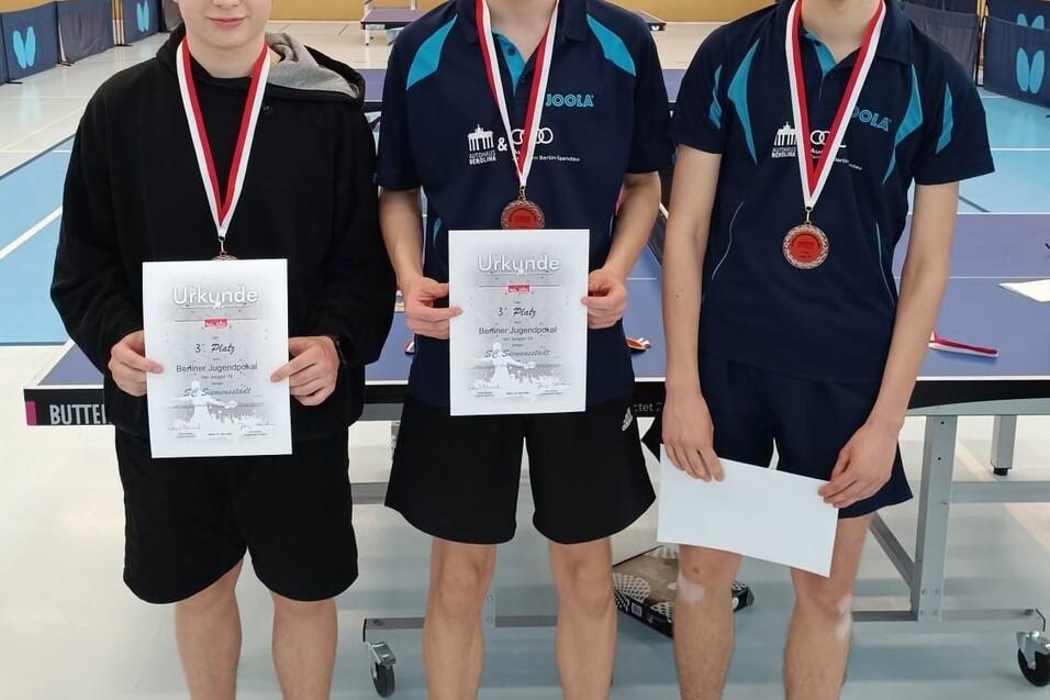 Drei Jugendliche mit Medaillen und Urkunden stehen vor einem Tischtennistisch in einer Sporthalle.