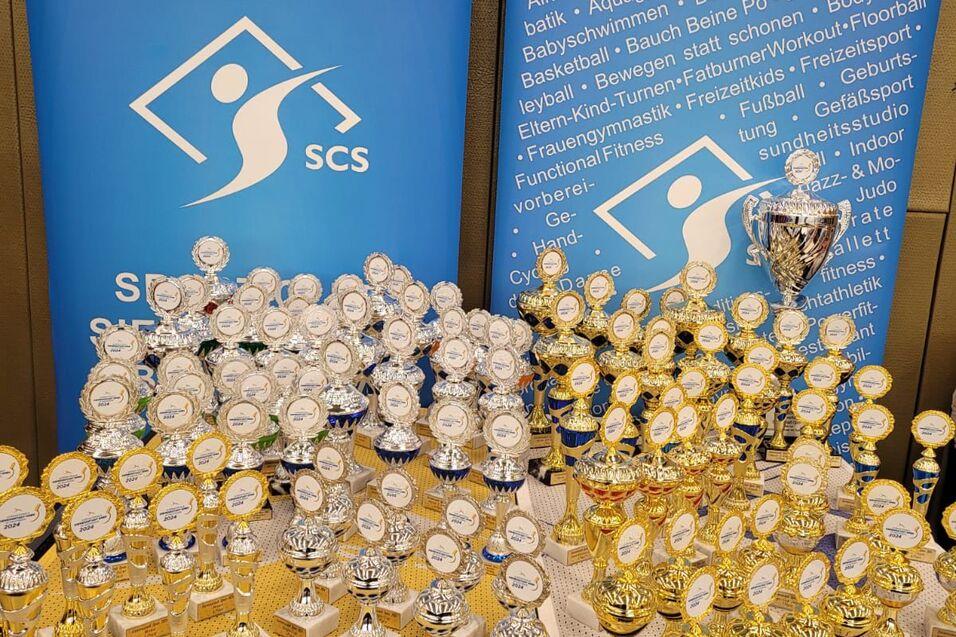 Eine Tischplatte mit zahlreichen Pokalen in Gold und Silber, umgeben von Bannern mit Sportinformationen.