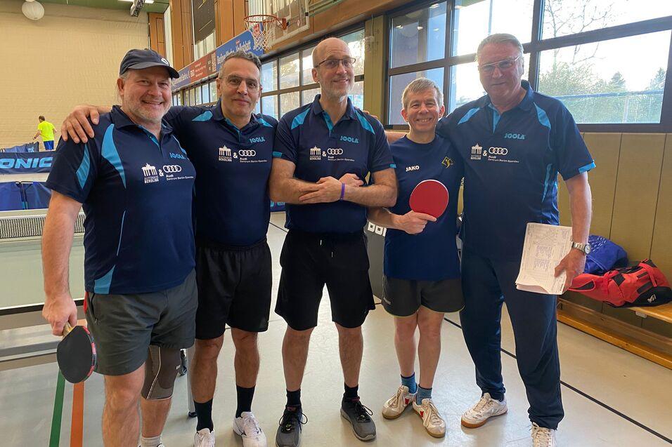 F&uuml;nf Tischtennisspieler in Uniformen stehen l&auml;chelnd in einer Sporthalle mit Tischtennisplatte im Hintergrund.