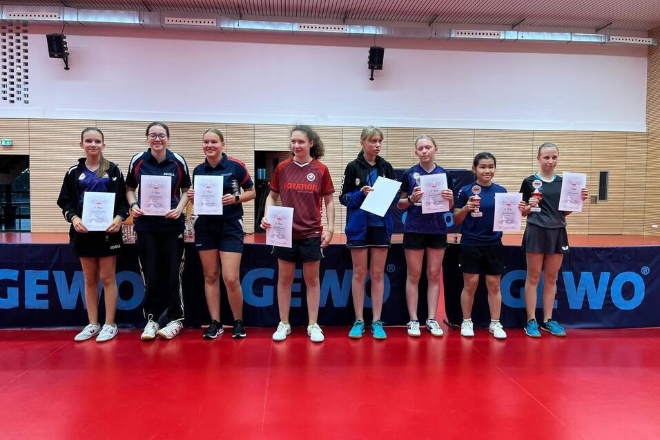 Acht Tischtennisspielerinnen stehen mit Urkunden und Pokalen vor einer Tischtennisplatte in einer Sporthalle.