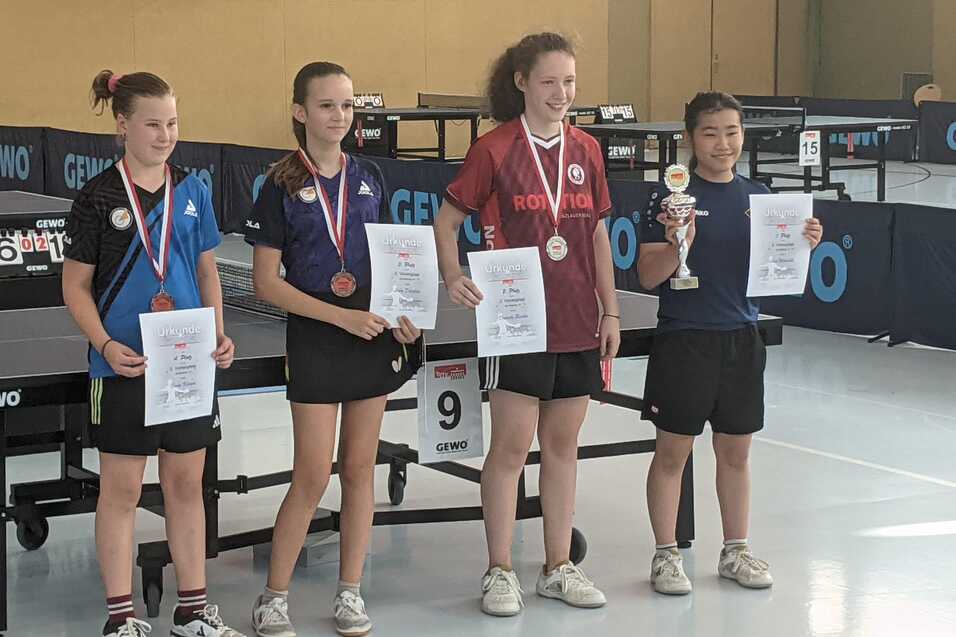 Vier Tischtennisspielerinnen halten Urkunden und Medaillen, stehen auf einem Platz mit Tischtennisplatten im Hintergrund.