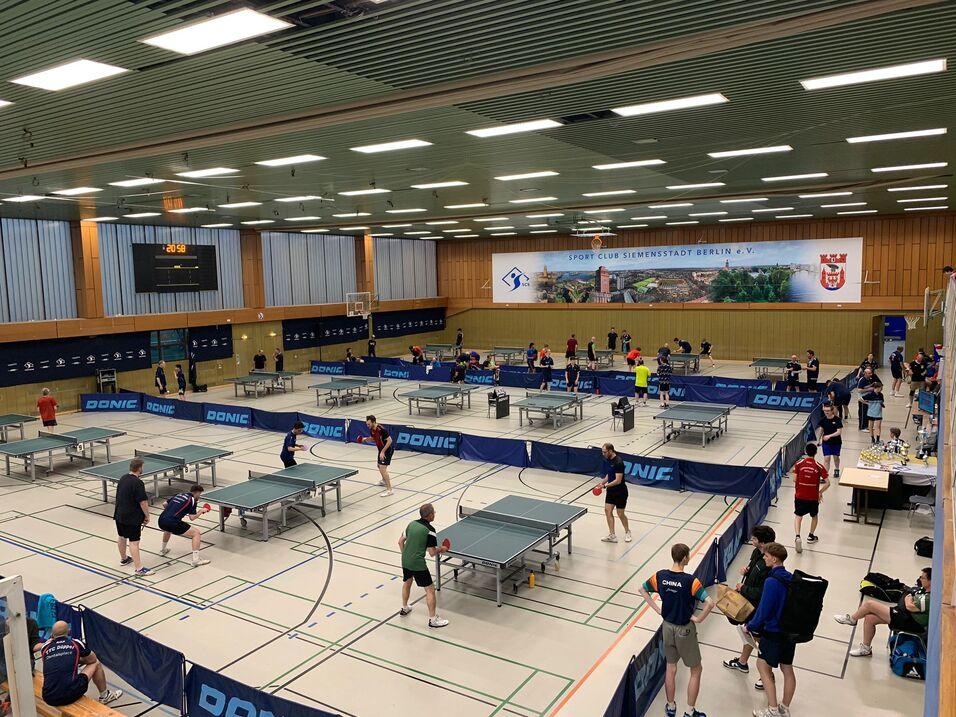 Tischtennisveranstaltung in einer Sporthalle mit mehreren Spielern an Tischen und einer digitalen Anzeigetafel im Hintergrund.