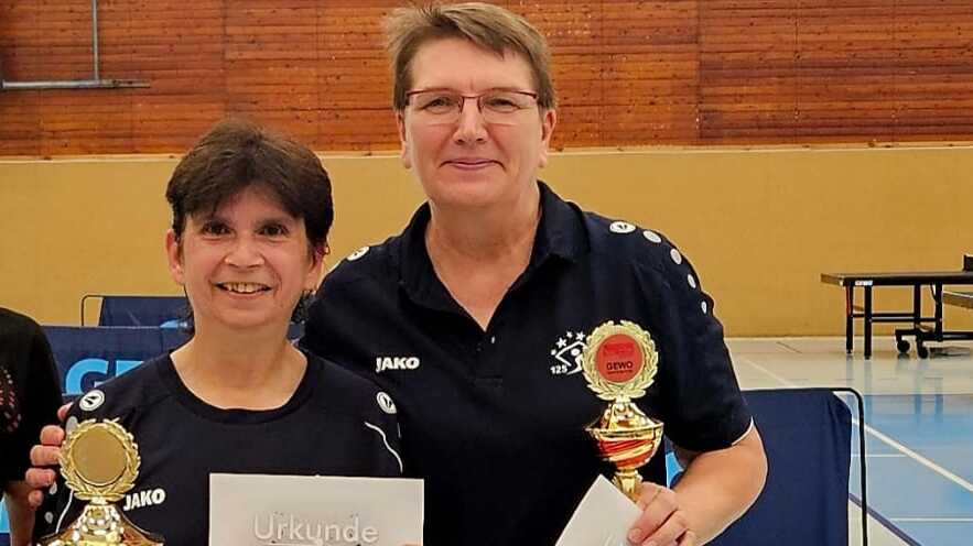 Zwei Tischtennisspielerinnen halten Pokale und Urkunden nach dem Turnier. Im Hintergrund sind Tischtennisplatten sichtbar.