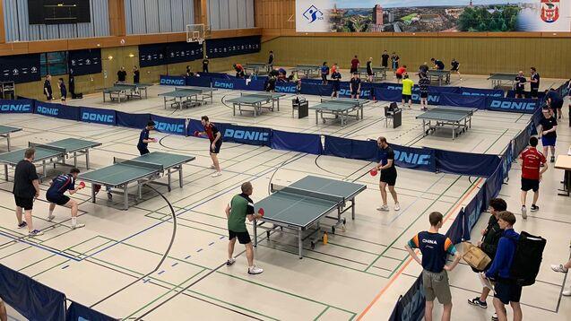 Tischtennisveranstaltung in einer Sporthalle mit mehreren Spielern an Tischen und einer digitalen Anzeigetafel im Hintergrund.