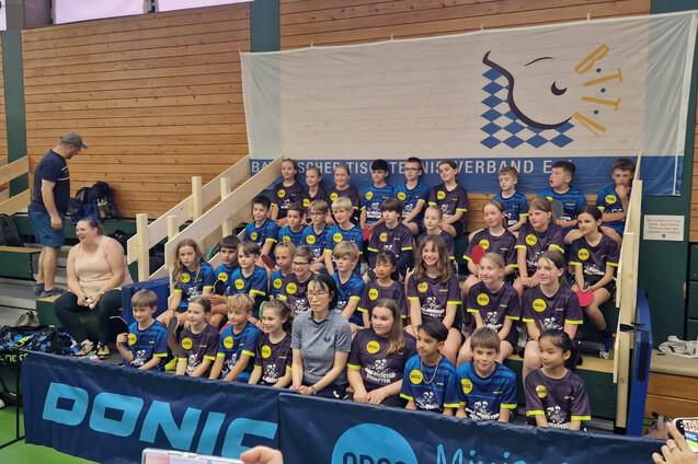 Gruppenfoto von Kindern in Sportkleidung mit Tischtennis-T-Shirts, versammelt vor einem Banner im Turnierumfeld.