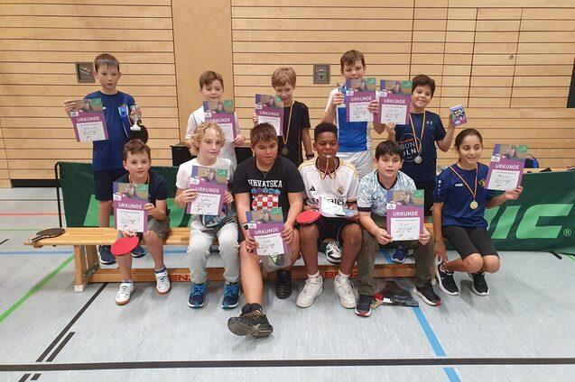 Gruppe von Kindern mit Urkunden und Medaillen nach einem Tischtennisturnier in einer Sporthalle.