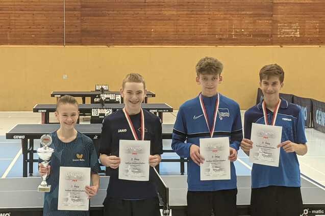 Vier Jugendliche stehen l&auml;chelnd vor Tischtennisplatten und halten Pokale sowie Urkunden in den H&auml;nden.