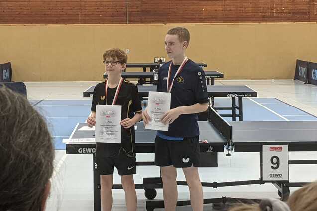 Zwei Tischtennisspieler stehen mit Urkunden und Medaillen auf einem Tischtennisturnier, Tischtennisplatten im Hintergrund.