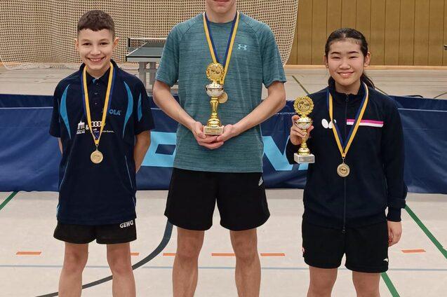 Drei Tischtennisspieler stehen mit Pokalen und Medaillen in einer Sporthalle, im Hintergrund ein Netz und Banner.