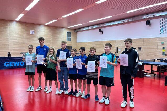 Gruppe von acht Jugendlichen h&auml;lt Urkunden und Pokale in einem Tischtennisraum mit rotem Boden.