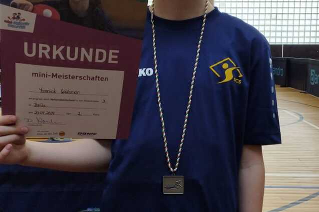 Junge mit Urkunde und Medaille f&uuml;r die mini-Meisterschaften im Tischtennis, stehend in einer Sporthalle.