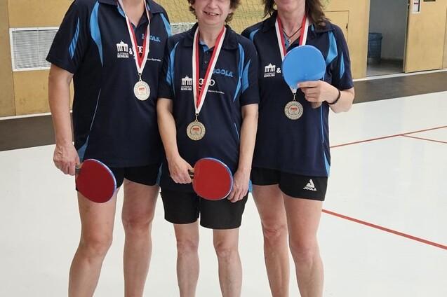 Drei Tischtennisspielerinnen in Sportkleidung halten Schl&auml;ger und Medaillen in einer Sporthalle.