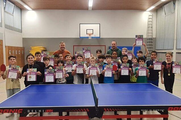 Kinder stehen in einer Sporthalle mit Urkunden und Medaillen vor einem Tischtennistisch.