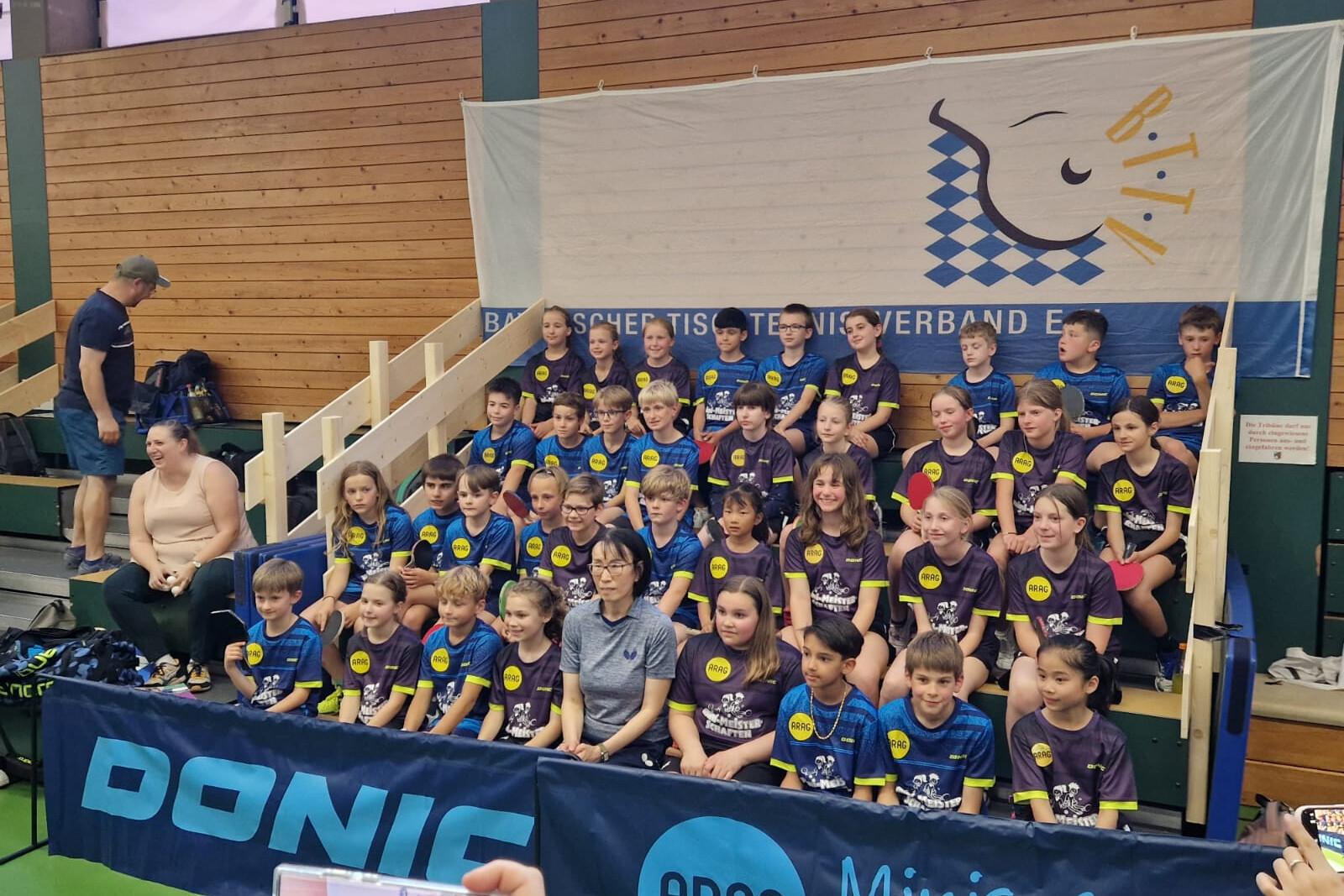 Gruppenfoto von Kindern in Sportkleidung mit Tischtennis-T-Shirts, versammelt vor einem Banner im Turnierumfeld.