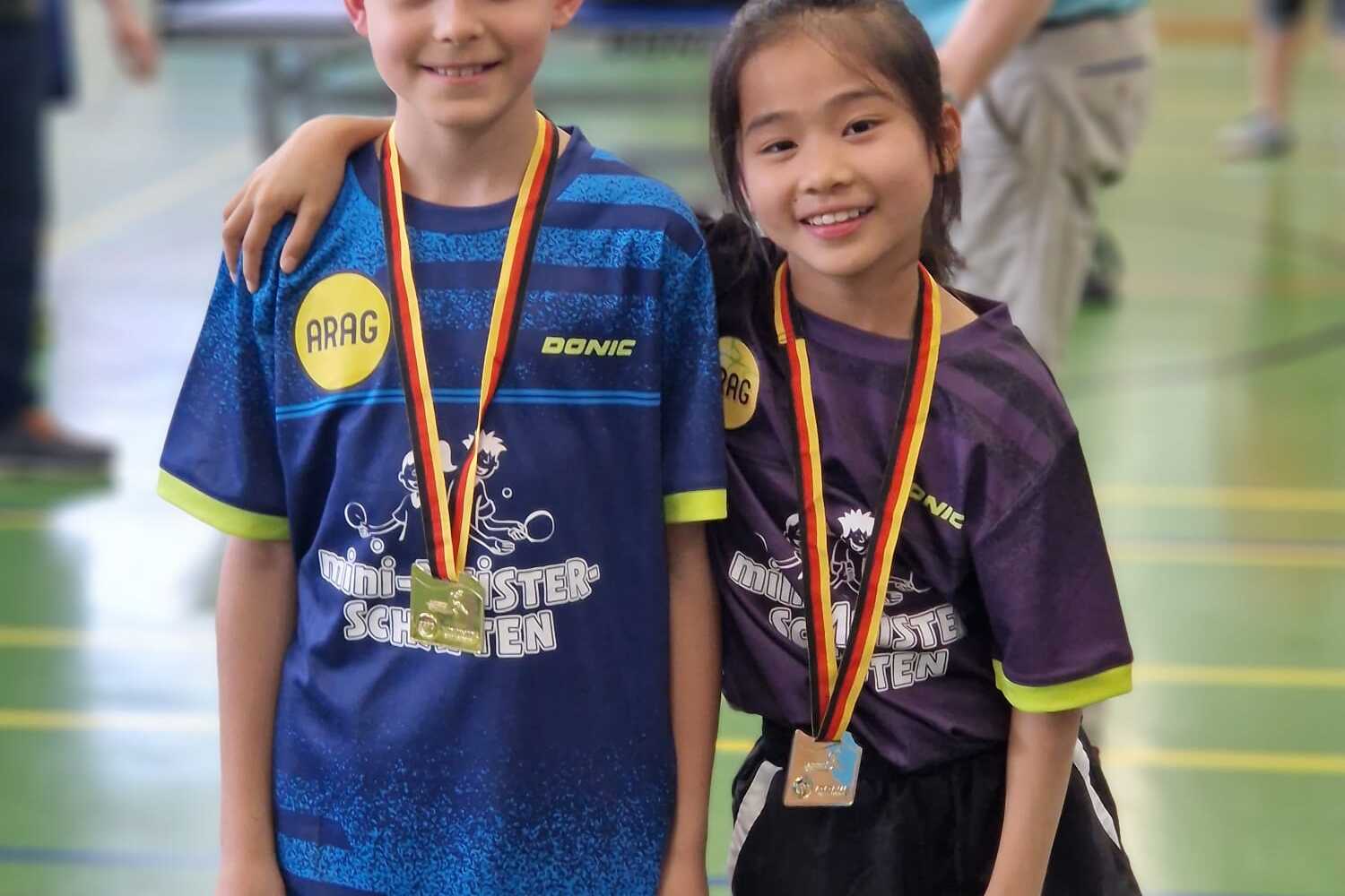 Zwei Kinder in Tischtennis-Trikots stehen l&auml;chelnd, Medaillen um den Hals, in einer Sporthalle.