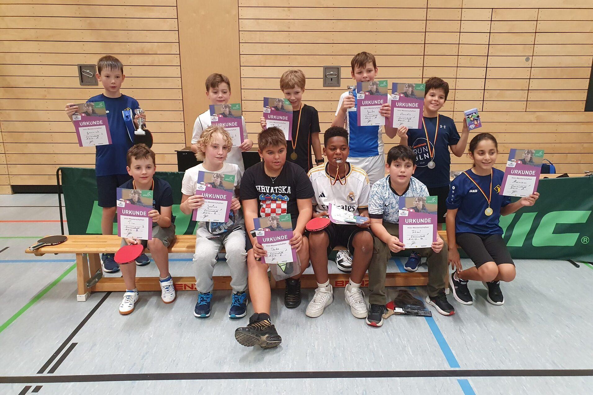 Gruppe von Kindern mit Urkunden und Medaillen nach einem Tischtennisturnier in einer Sporthalle.