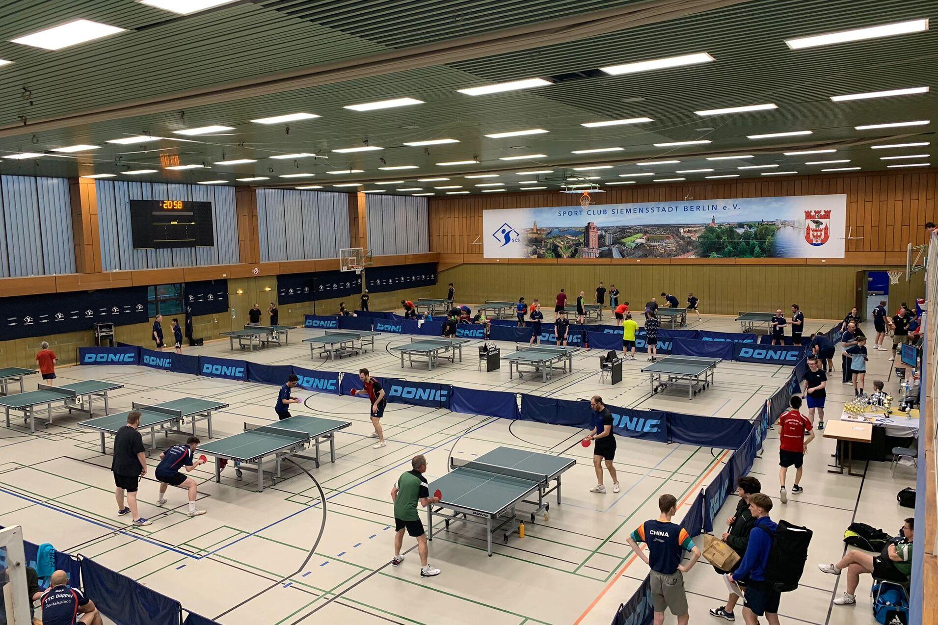 Tischtennisveranstaltung in einer Sporthalle mit mehreren Spielern an Tischen und einer digitalen Anzeigetafel im Hintergrund.