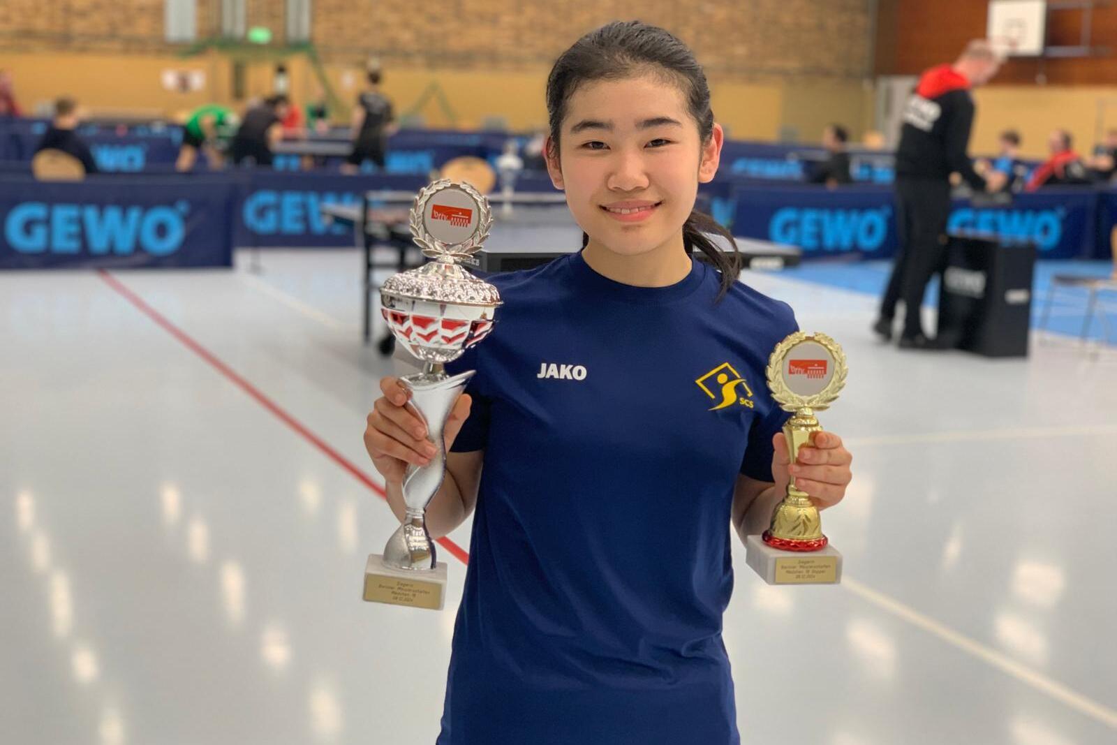 Junge Sportlerin h&auml;lt zwei Troph&auml;en in der Hand, im Hintergrund eine Tischtennis-Spielhalle mit Spielern.
