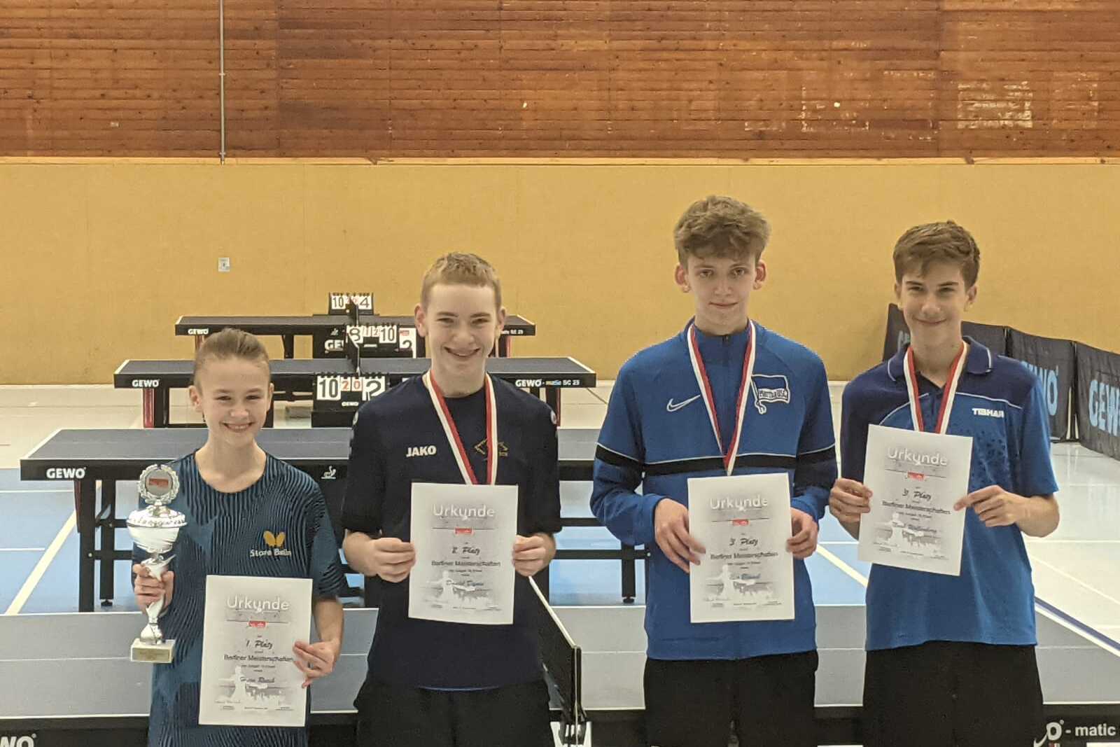 Vier Jugendliche stehen l&auml;chelnd vor Tischtennisplatten und halten Pokale sowie Urkunden in den H&auml;nden.