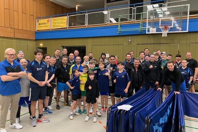 Gruppenfoto von Tischtennisspielern unterschiedlicher Altersgruppen in einer Sporthalle, l&auml;chelnd und Daumen hoch.