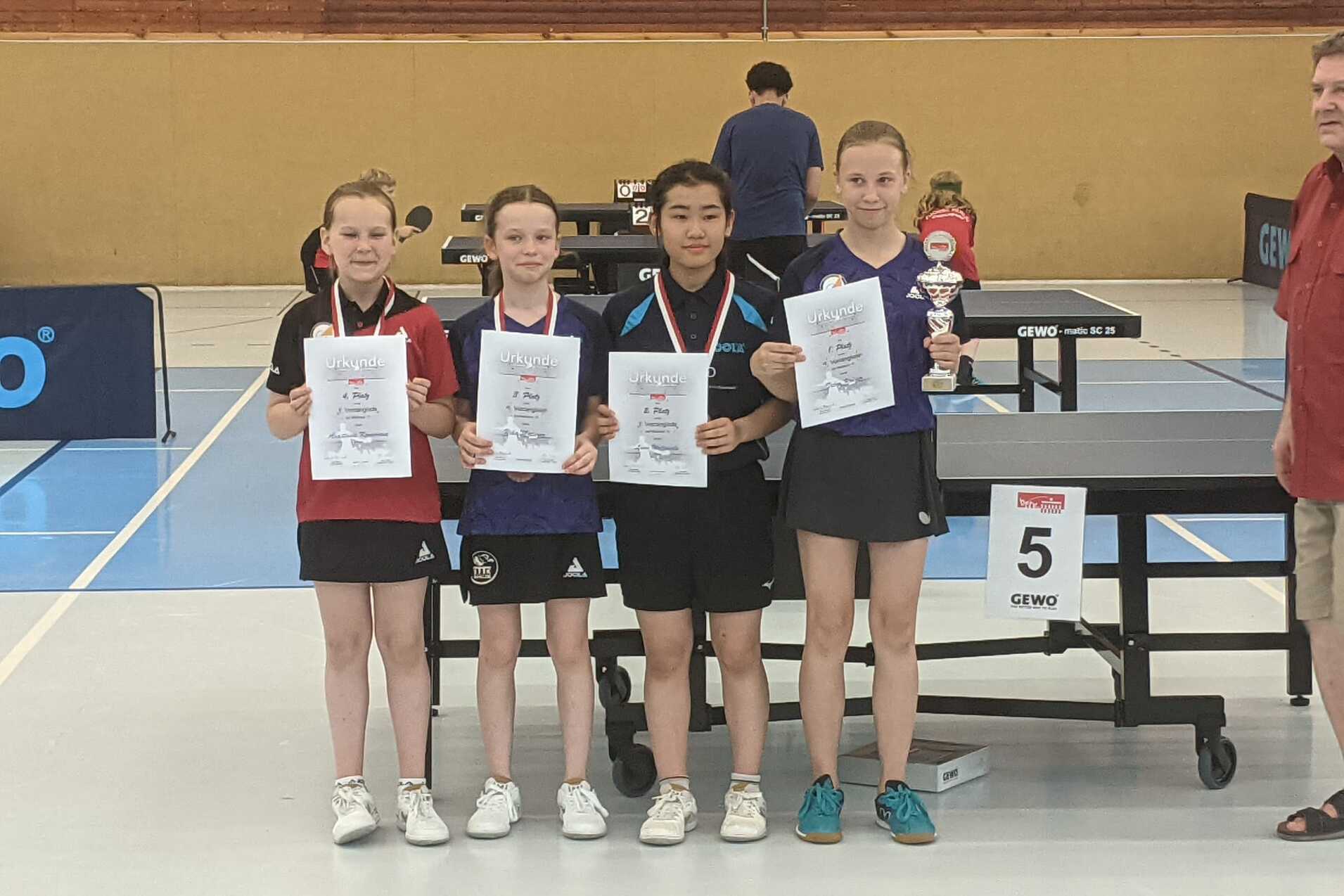 Vier M&auml;dchen stehen in Sportkleidung vor einem Tischtennistisch, halten Urkunden und einen Pokal.