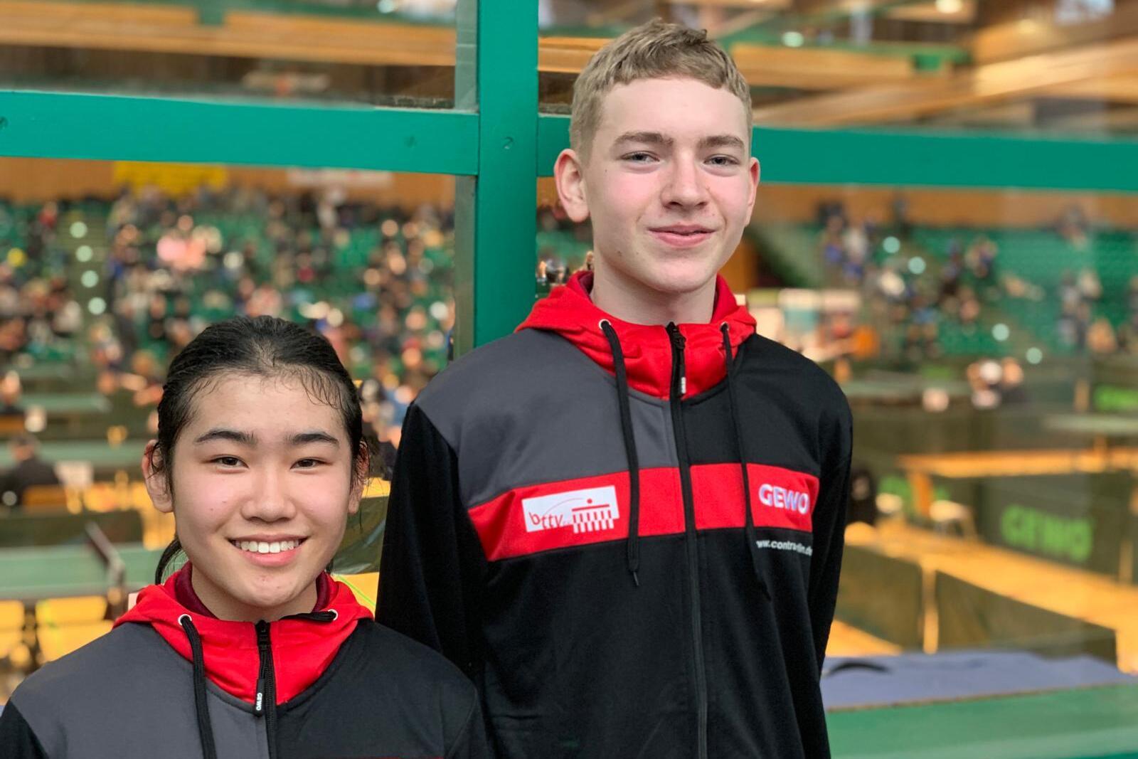 Zwei Jugendliche in roten und schwarzen Sportjacken stehen vor einer Tischtennishalle mit Zuschauern im Hintergrund.