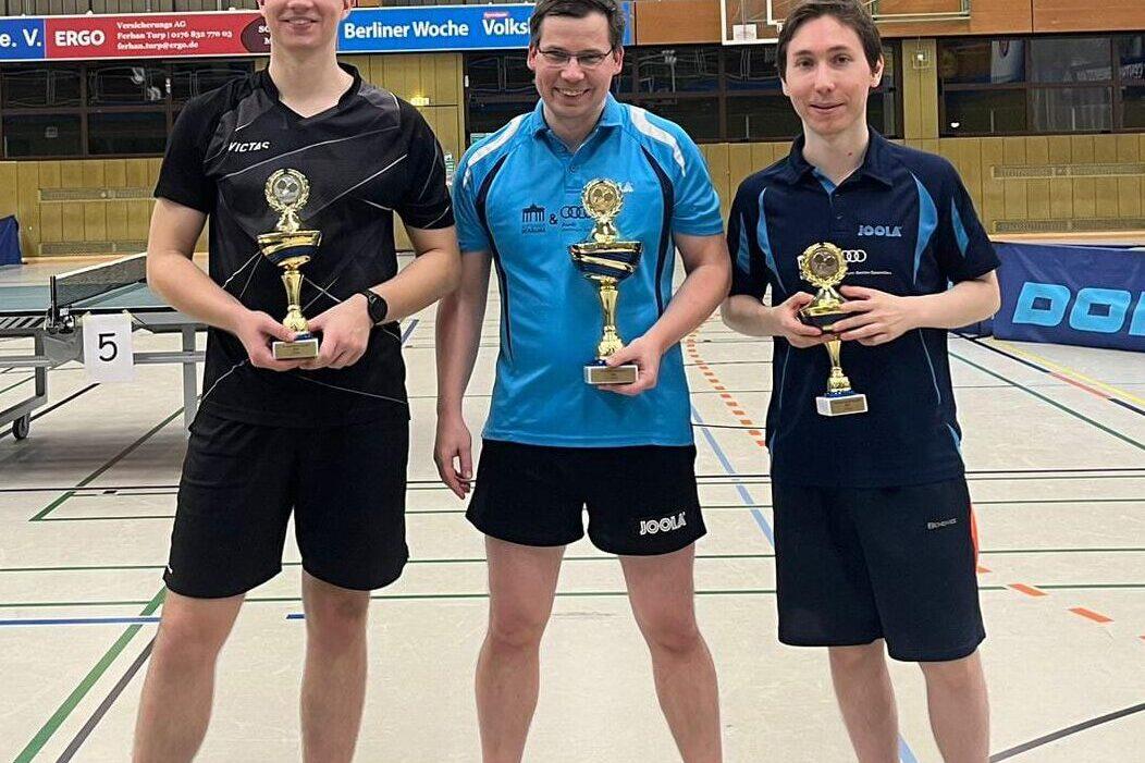 Drei Tischtennisspieler stehen in Sportkleidung nebeneinander und halten ihre Pokale in einer Sporthalle.