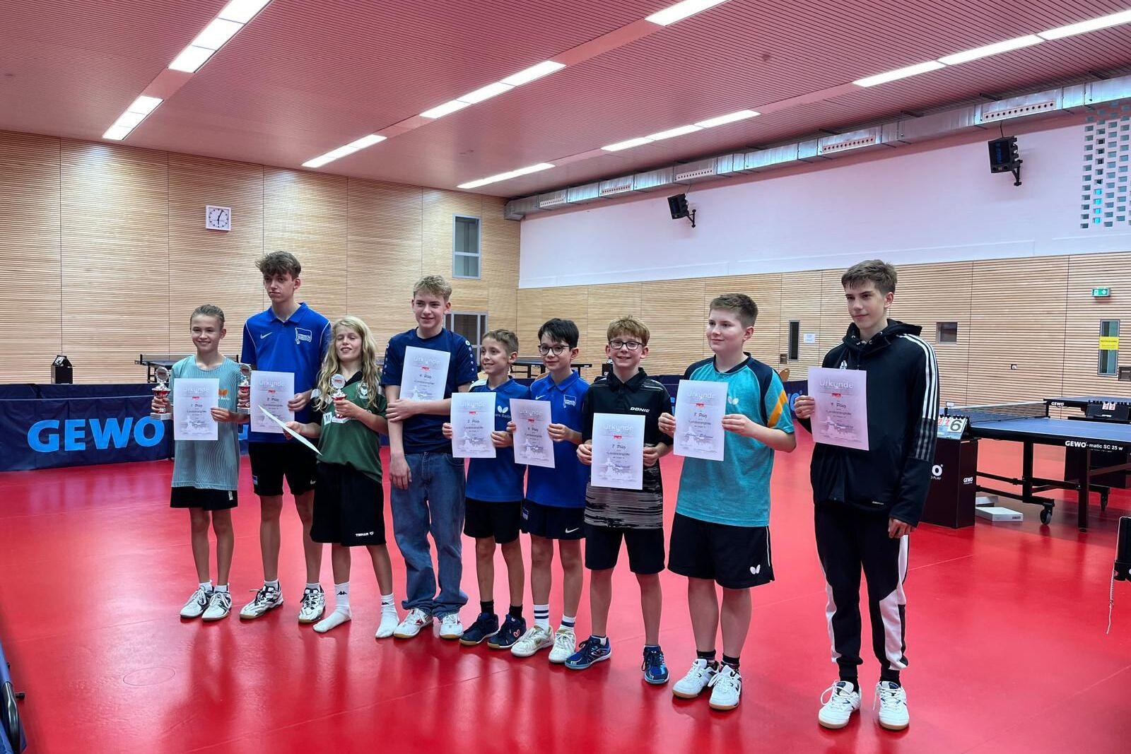 Gruppe von acht Jugendlichen hält Urkunden und Pokale in einem Tischtennisraum mit rotem Boden.