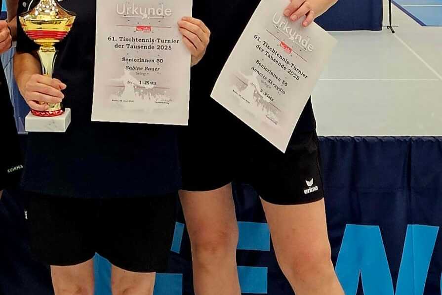 Zwei Tischtennisspielerinnen halten Pokale und Urkunden nach dem Turnier. Im Hintergrund sind Tischtennisplatten sichtbar.