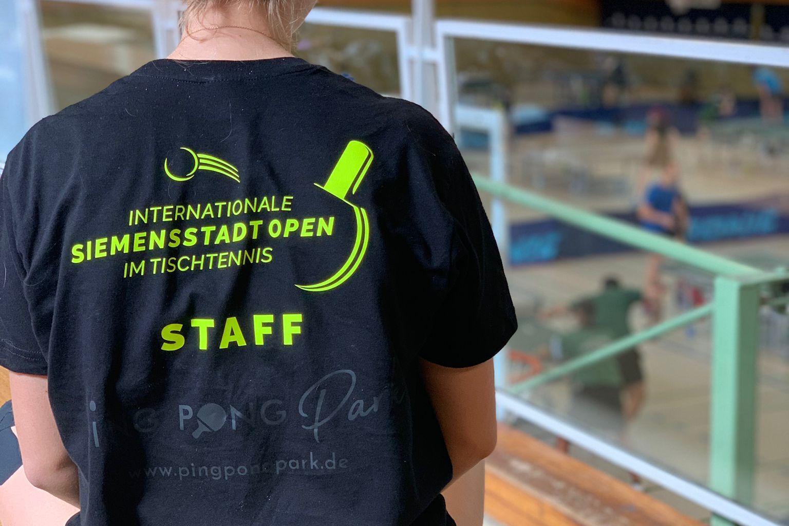 R&uuml;ckansicht einer Person in schwarzem Shirt mit Aufdruck &bdquo;Internationale Siemenstadt Open im Tischtennis STAFF&ldquo;.
