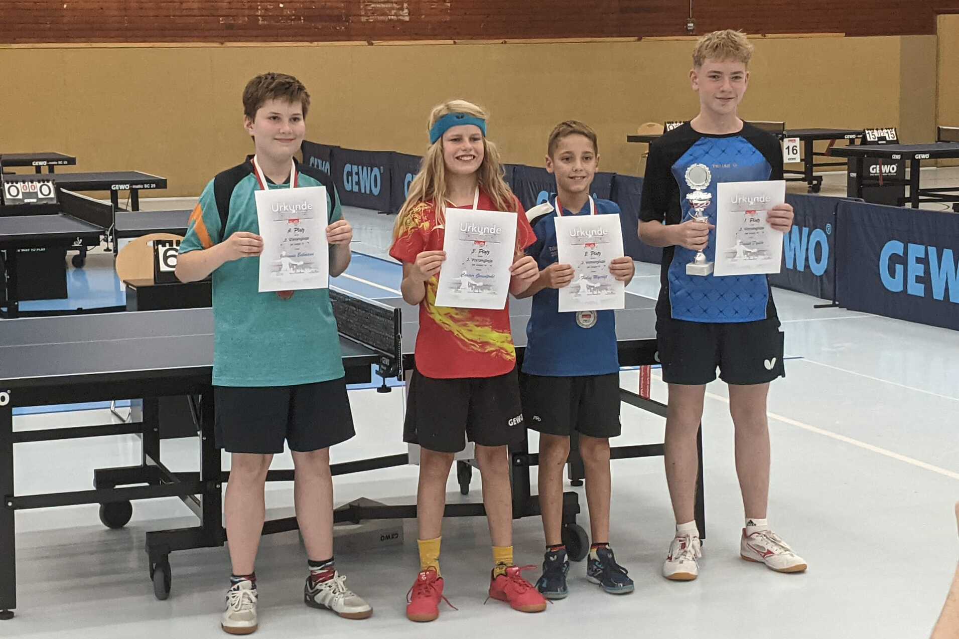 Vier Tischtennisspieler halten Urkunden und Pokale in einer Turnierhalle mit Tischtennisplatten im Hintergrund.
