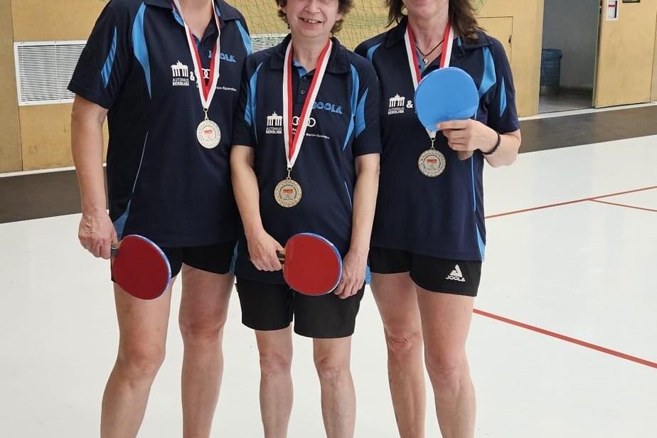 Drei Tischtennisspielerinnen in Sportkleidung halten Schl&auml;ger und Medaillen in einer Sporthalle.