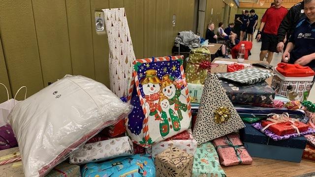 Bunt verpackte Geschenkpäckchen und -tüten auf einem Tisch in einer Halle, im Hintergrund sind Menschen zu sehen.