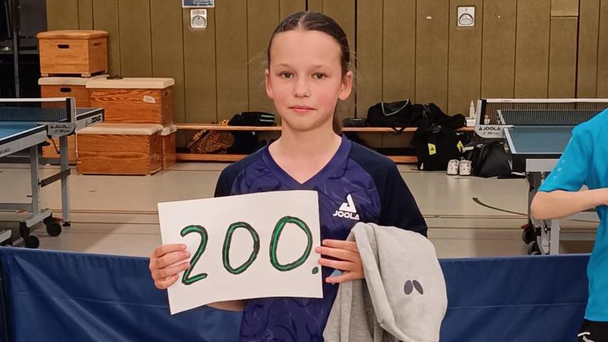 Mädchen hält ein Plakat mit der Zahl „200“ in einer Sporthalle, während ein weiterer Junge im Hintergrund steht.