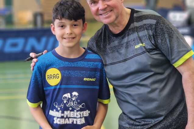 Porträt eines Jungen mit Tischtennisschläger und einem Mann in Sportkleidung, Hintergrund: Tischtennishalle.