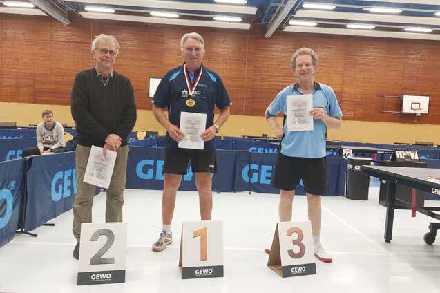 Drei Männer in Sportkleidung stehen mit Urkunden und Medaillen vor einem Tischtennis-Tisch. Anzeigetafeln zeigen die Plätze 1, 2 und 3.