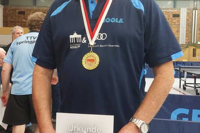Mann mit Brille hält eine Urkunde und Medaille nach Tischtennisturnier, steht in einer Sporthalle.