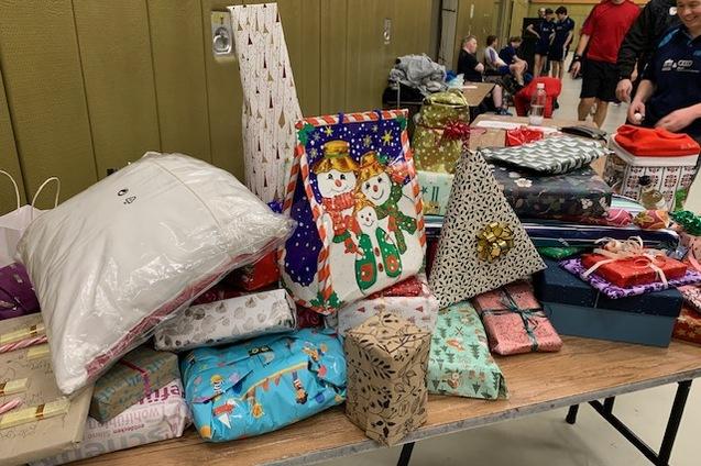 Bunt verpackte Geschenkpäckchen und -tüten auf einem Tisch in einer Halle, im Hintergrund sind Menschen zu sehen.