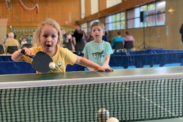 Ein Mädchen mit blonden Locken spielt Tischtennis, während ein Junge im Hintergrund zuschaut.