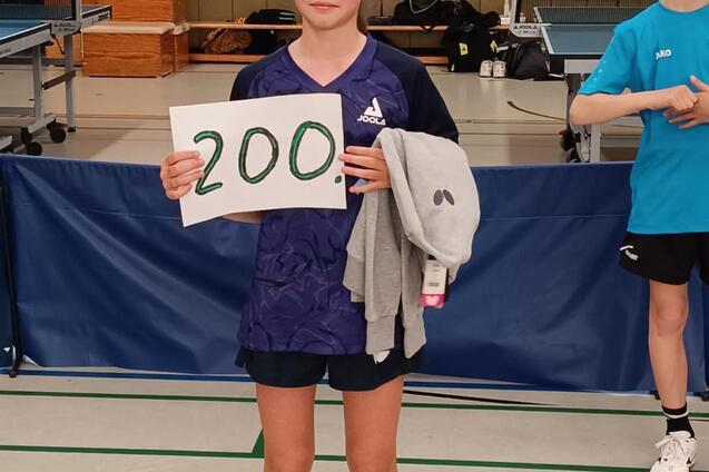 Mädchen hält ein Plakat mit der Zahl „200“ in einer Sporthalle, während ein weiterer Junge im Hintergrund steht.