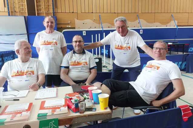 F&uuml;nf Personen in einheitlichen Shirts sitzen an einem Tisch mit Getr&auml;nken und Unterlagen in einer Sporthalle.