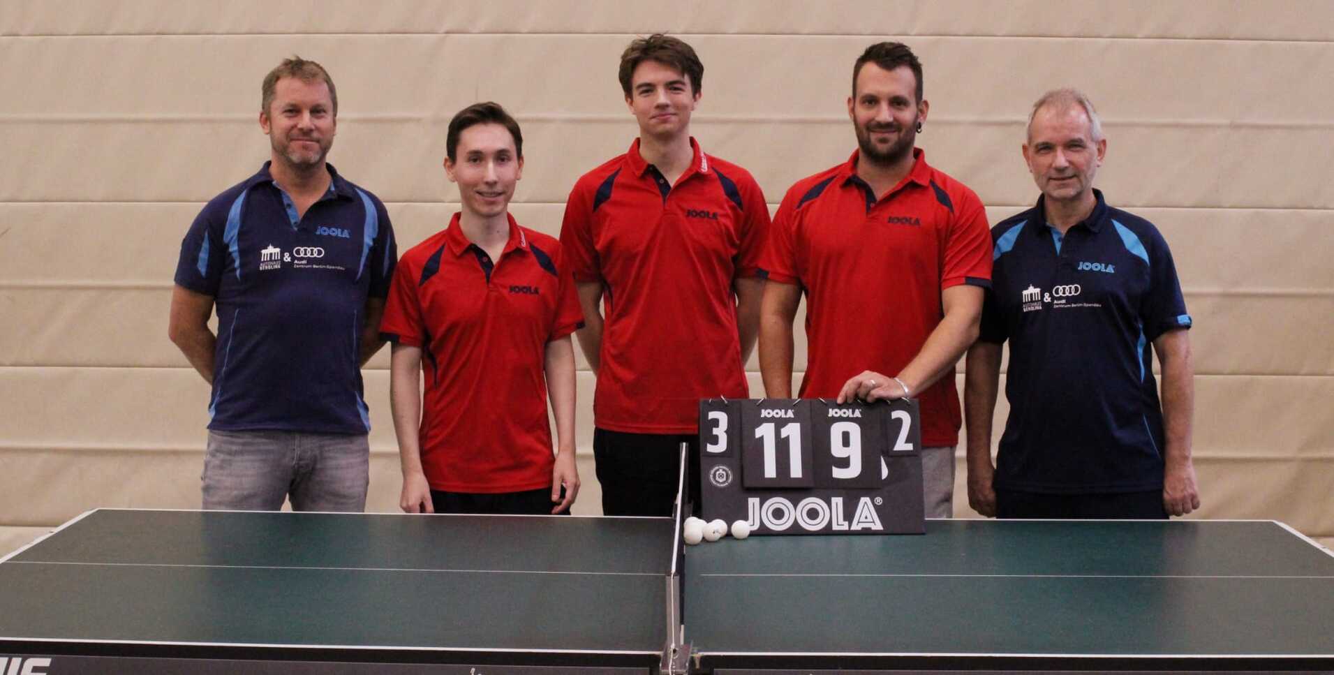 Fünf Tischtennisspieler in roten und blauen jerseys stehen vor einem Tisch mit Scoreboard und Tischtennisbällen.